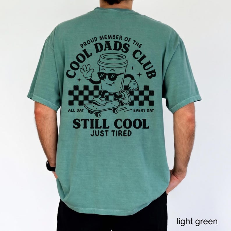 Pu&ograve; includere: T-shirt verde chiaro con la scritta "PROUD MEMBER OF THE COOL DADS CLUB" e una tazza di caff&egrave; dei cartoni animati su uno skateboard. La maglietta dice anche "ALL DAY EVERY DAY STILL COOL JUST TIRED".
