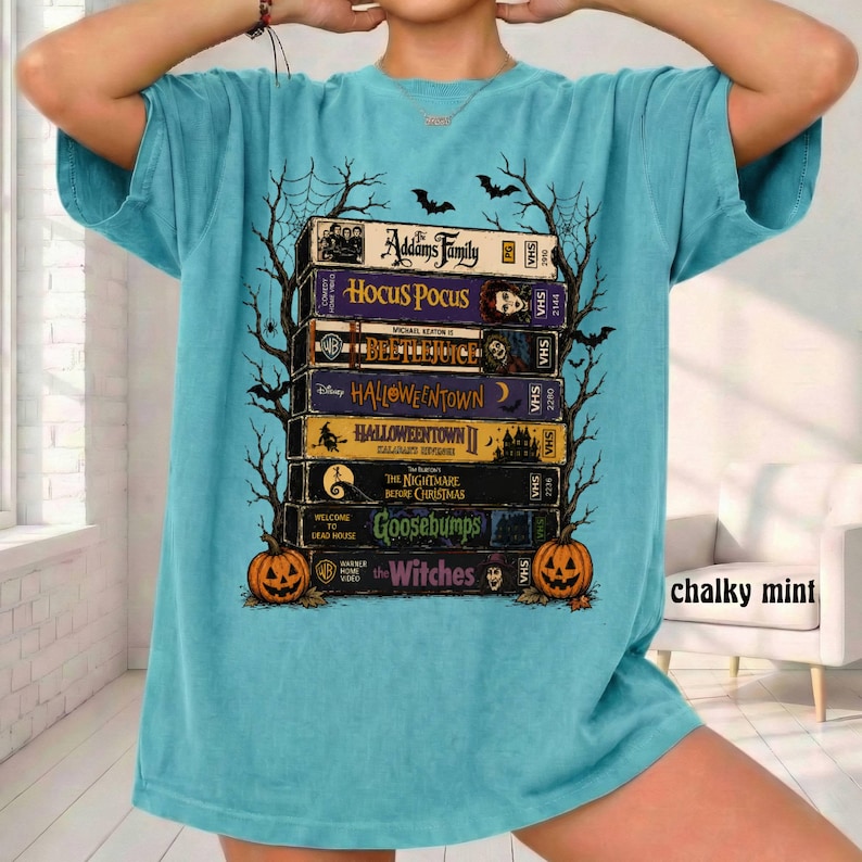 Op de afbeelding: Een blauwgroen t-shirt met een afbeelding van een stapel VHS-banden met titels zoals Addams Family, Hocus Pocus, Beetlejuice, Halloweentown, Halloweentown II, The Nightmare Before Christmas, Goosebumps en The Witches, omlijst door zwarte boomsilhouetten.
