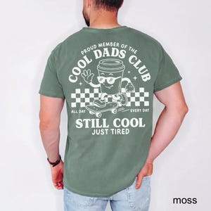 Pu&ograve; includere: T-shirt verde muschio con grafica sul retro con la scritta "COOL DADS CLUB" con un personaggio a forma di tazza di caff&egrave; sullo skateboard. Il testo "ALL DAY EVERY DAY STILL COOL JUST TIRED" &egrave; stampato sulla maglietta.