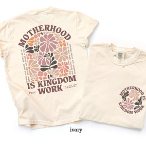 May include: Two ivory-coloured t-shirts. One has a floral design and the text "MOTHERHOOD IS KINGDOM WORK" on the back. The other has the same design on the front. The shirts are made of a soft material.