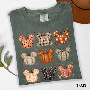 May include: A moss green t-shirt featuring a graphic print of nine pumpkins, each shaped like mouse ears with distinct patterns including leopard print, buffalo plaid, floral, polka dots, and solid orange, accented by small hearts and leaves.