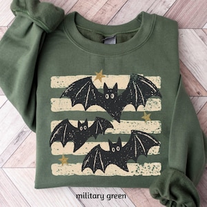 May include: An olive green crewneck sweatshirt featuring a graphic print of three black bats flying across four horizontal cream-colored distressed stripes. The design includes small gold-colored stars scattered between the bats and stripes.