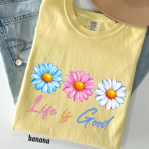 Pode incluir: Uma camiseta amarela com tr&ecirc;s flores de margarida em azul, rosa e branco, com centros laranja. A camisa tem o texto "Life is Good" em script rosa e azul, e a palavra "banana" na parte inferior.