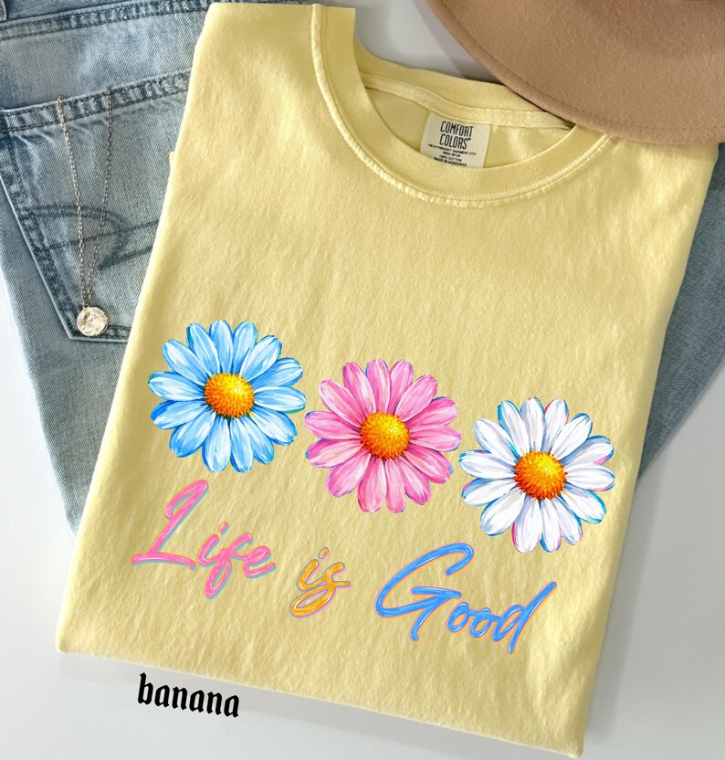 Pode incluir: Uma camiseta amarela com tr&ecirc;s flores de margarida em azul, rosa e branco, com centros laranja. A camisa tem o texto "Life is Good" em script rosa e azul, e a palavra "banana" na parte inferior.