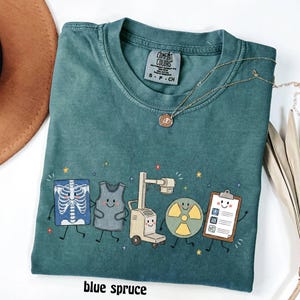 May include: A blue spruce colored t-shirt featuring a graphic print of five cartoon medical characters including an x-ray, a lead apron, an imaging machine, a radiation symbol, and a clipboard, all with smiling faces and small colorful stars.