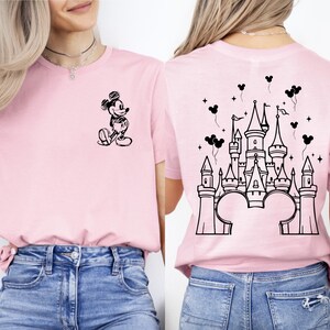 Puede incluir: Camiseta rosa claro con un boceto negro de Mickey Mouse en la parte delantera y un dise&ntilde;o de castillo en la parte trasera. El dise&ntilde;o del castillo incluye orejas de Mickey Mouse y globos.