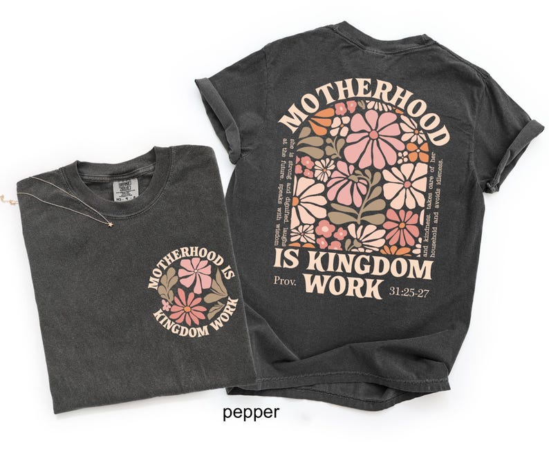 May include: Two dark grey t-shirts. One features a floral design with the text "MOTHERHOOD IS KINGDOM WORK" and Bible verse 31:25-27. The other shirt has a smaller version of the design on the front. The shirts are made of a soft material.