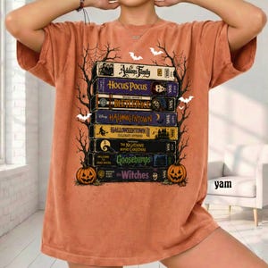 May include: An orange t-shirt featuring a graphic print of a stack of VHS tapes titled The Addams Family, Hocus Pocus, Beetlejuice, Halloweentown, Halloweentown II, The Nightmare Before Christmas, Goosebumps, and The Witches, framed by black spiderwebs.