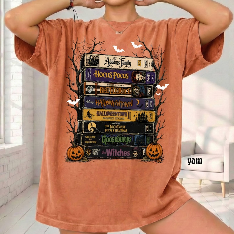 Op de afbeelding: Een oranje t-shirt met een grafische print van een stapel VHS-banden met de titels The Addams Family, Hocus Pocus, Beetlejuice, Halloweentown, Halloweentown II, The Nightmare Before Christmas, Goosebumps en The Witches, omlijst door zwarte spinnenwebben.