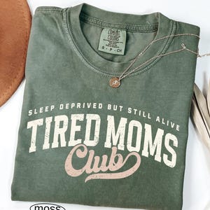 May include: A moss green t-shirt with the text "SLEEP DEPRIVED BUT STILL ALIVE TIRED MOMS Club" in white and pink lettering. A gold-colored necklace with a small pendant rests on the shirt. The shirt is folded on a white surface.