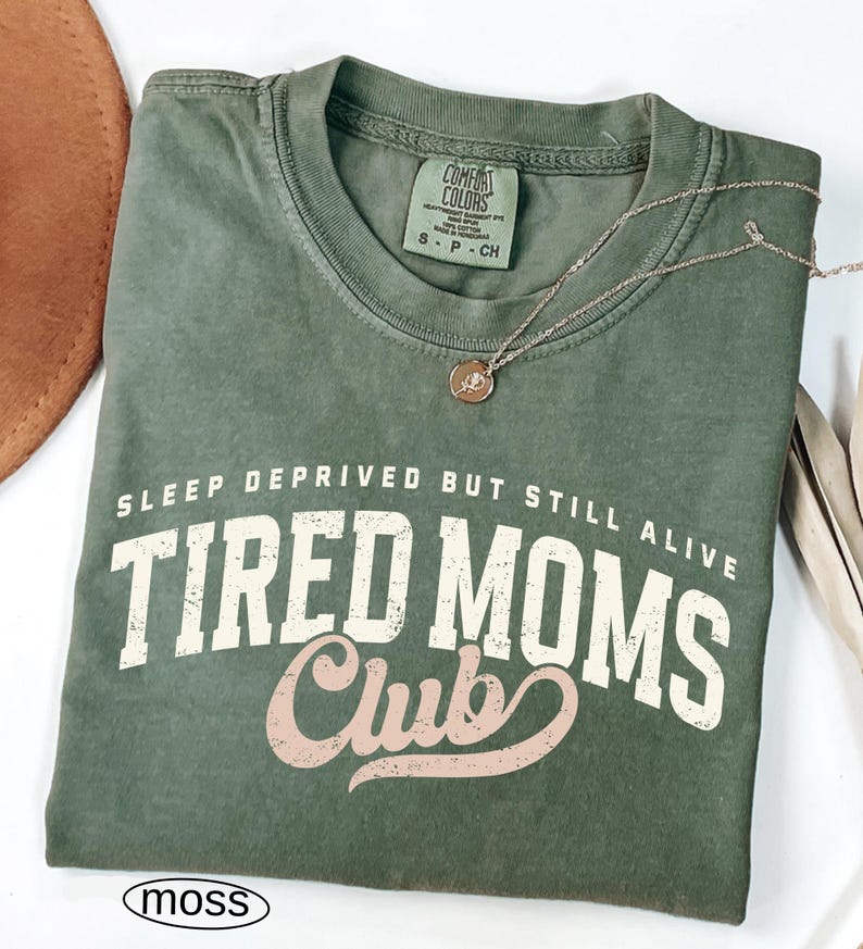 May include: A moss green t-shirt with the text "SLEEP DEPRIVED BUT STILL ALIVE TIRED MOMS Club" in white and pink lettering. A gold-colored necklace with a small pendant rests on the shirt. The shirt is folded on a white surface.