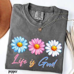 Pode incluir: Uma camiseta cinza Comfort Colors com tr&ecirc;s flores de margarida em azul, rosa e branco, cada uma com um centro amarelo. O texto "Life is Good" est&aacute; abaixo das flores em uma escrita colorida. Um colar de prata com um pingente est&aacute; na camiseta.