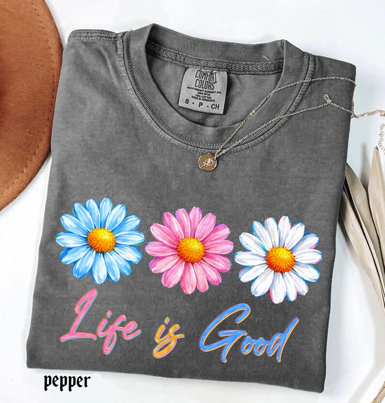 Pode incluir: Uma camiseta cinza Comfort Colors com tr&ecirc;s flores de margarida em azul, rosa e branco, cada uma com um centro amarelo. O texto "Life is Good" est&aacute; abaixo das flores em uma escrita colorida. Um colar de prata com um pingente est&aacute; na camiseta.