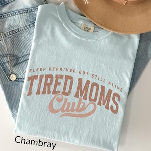 May include: A light blue t-shirt with the text "SLEEP DEPRIVED BUT STILL ALIVE TIRED MOMS Club" in a distressed, brown font. The word "Chambray" is printed in black at the bottom left. The shirt is folded on top of blue jeans.