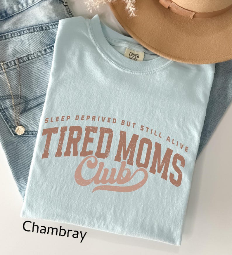 May include: A light blue t-shirt with the text "SLEEP DEPRIVED BUT STILL ALIVE TIRED MOMS Club" in a distressed, brown font. The word "Chambray" is printed in black at the bottom left. The shirt is folded on top of blue jeans.