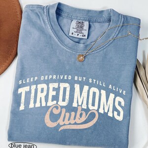 May include: A blue jean Comfort Colors t-shirt with the text "SLEEP DEPRIVED BUT STILL ALIVE TIRED MOMS Club" in white and pink lettering. A gold necklace with a pendant rests on the shirt.