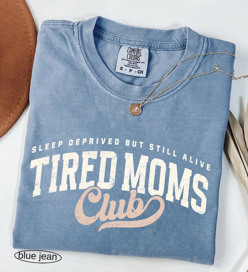 May include: A blue jean Comfort Colors t-shirt with the text "SLEEP DEPRIVED BUT STILL ALIVE TIRED MOMS Club" in white and pink lettering. A gold necklace with a pendant rests on the shirt.