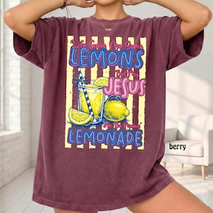 May include: Berry-colored t-shirt with a graphic of a lemonade drink, lemons, and the text "Life Gave Me Some Lemons But My Jesus He Be Makin' Lemonade." The shirt has a relaxed fit.