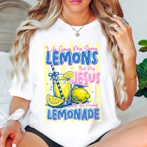 May include: White t-shirt with a colorful graphic that reads "Life Gave Me Some Lemons But My Jesus He Be Makin' Lemonade." The design includes a glass of lemonade, lemons, and a striped straw. The text is in blue and pink.