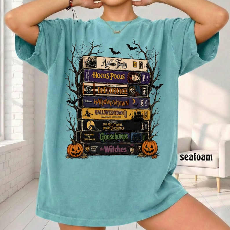 Op de afbeelding: Een lichtblauw t-shirt met korte mouwen en een afbeelding van gestapelde VHS-banden met titels als The Addams Family, Hocus Pocus, Beetlejuice, Halloweentown, The Nightmare Before Christmas, Goosebumps en The Witches, omlijst door zwarte boom-silhouetten.