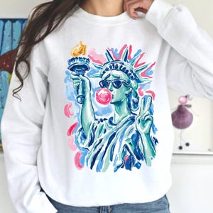 May include: A white long-sleeve sweatshirt featuring a colorful graphic print of the Statue of Liberty wearing dark sunglasses and blowing a bright pink bubblegum bubble, while holding a torch and making a peace sign with the other hand.
