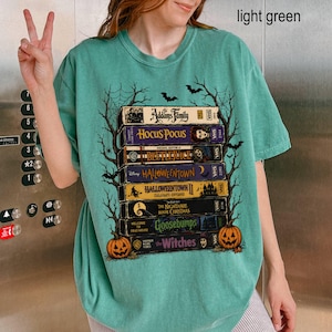 Op de afbeelding: Een lichtgroen t-shirt met een grafische print van een stapel vintage VHS-banden met titels zoals The Addams Family, Hocus Pocus, Beetlejuice, Halloweentown, The Nightmare Before Christmas, Goosebumps en The Witches, omlijst door boomtakken.