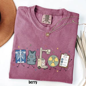May include: A berry-colored t-shirt featuring a graphic print of five cartoon medical characters: a smiling chest x-ray, a lead apron, an x-ray machine, a radiation symbol, and a clipboard. The shirt has a Comfort Colors brand label at the neck.