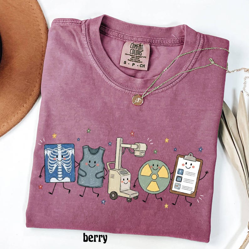 May include: A berry-colored t-shirt featuring a graphic print of five cartoon medical characters: a smiling chest x-ray, a lead apron, an x-ray machine, a radiation symbol, and a clipboard. The shirt has a Comfort Colors brand label at the neck.