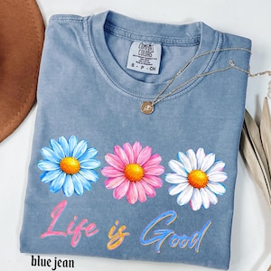 Pode incluir: Camiseta azul jeans com tr&ecirc;s flores de margarida em azul, rosa e branco com centros amarelos. As palavras "Life is Good" est&atilde;o escritas em um script colorido na frente. Um colar de ouro com um pequeno pingente est&aacute; sobre a camisa.