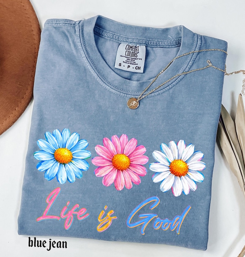 Pode incluir: Camiseta azul jeans com tr&ecirc;s flores de margarida em azul, rosa e branco com centros amarelos. As palavras "Life is Good" est&atilde;o escritas em um script colorido na frente. Um colar de ouro com um pequeno pingente est&aacute; sobre a camisa.