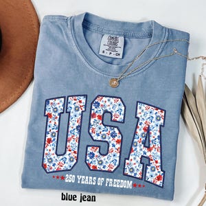 May include: A blue jean Comfort Colors t-shirt with the word "USA" in large, floral-patterned letters. The shirt also features the text "250 YEARS OF FREEDOM" and a gold necklace. The shirt is a size small.