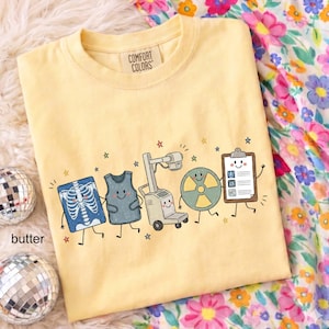 May include: A butter yellow t-shirt with a Comfort Colors label at the neck. The graphic features five cartoon characters: an X-ray of a ribcage, a lead apron, an X-ray machine, a radiation symbol, and a clipboard, all surrounded by small stars.