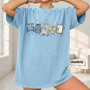 May include: A light blue t-shirt featuring a graphic print of five anthropomorphic medical imaging items, including an X-ray, a lead apron, an X-ray machine, a radiation symbol, and a clipboard, all decorated with small colorful stars.