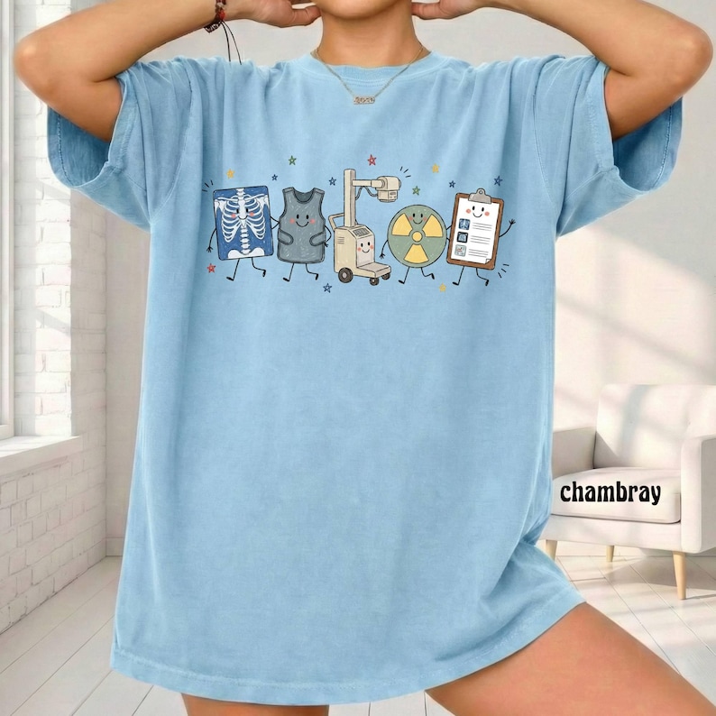 May include: A light blue t-shirt featuring a graphic print of five anthropomorphic medical imaging items, including an X-ray, a lead apron, an X-ray machine, a radiation symbol, and a clipboard, all decorated with small colorful stars.