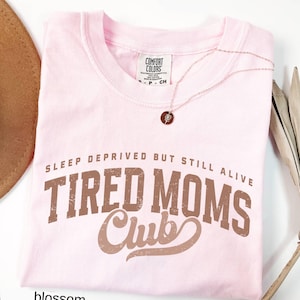May include: A light pink t-shirt with the text "SLEEP DEPRIVED BUT STILL ALIVE TIRED MOMS Club" in brown lettering. A delicate necklace with a small pendant rests on the shirt. The shirt is folded and has a tag that says "Comfort Colors".