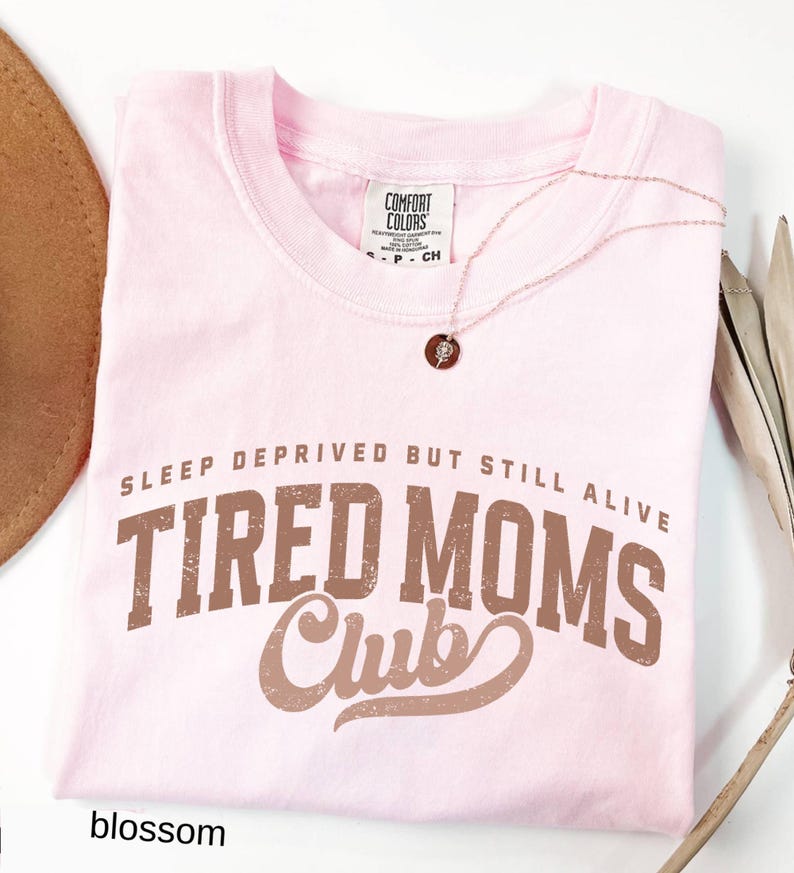 May include: A light pink t-shirt with the text "SLEEP DEPRIVED BUT STILL ALIVE TIRED MOMS Club" in brown lettering. A delicate necklace with a small pendant rests on the shirt. The shirt is folded and has a tag that says "Comfort Colors".