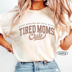 May include: Ivory-colored t-shirt with the text "SLEEP DEPRIVED BUT STILL ALIVE TIRED MOMS Club" in brown lettering. The shirt is paired with blue denim jeans. The word "ivory" is printed on the bottom right.