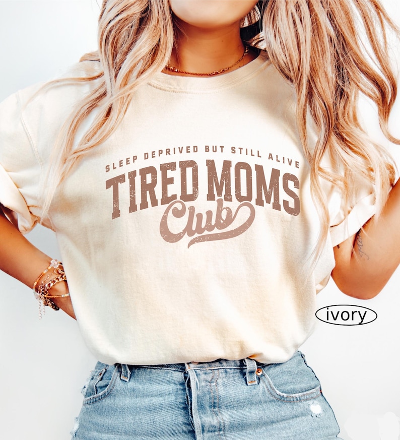 May include: Ivory-colored t-shirt with the text "SLEEP DEPRIVED BUT STILL ALIVE TIRED MOMS Club" in brown lettering. The shirt is paired with blue denim jeans. The word "ivory" is printed on the bottom right.