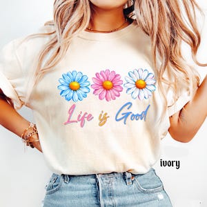 Pode incluir: Camiseta cor marfim com tr&ecirc;s margaridas azuis, rosas e brancas com centros amarelos. A frase "Life is Good" est&aacute; escrita abaixo das flores em um gradiente de rosa, amarelo e azul.