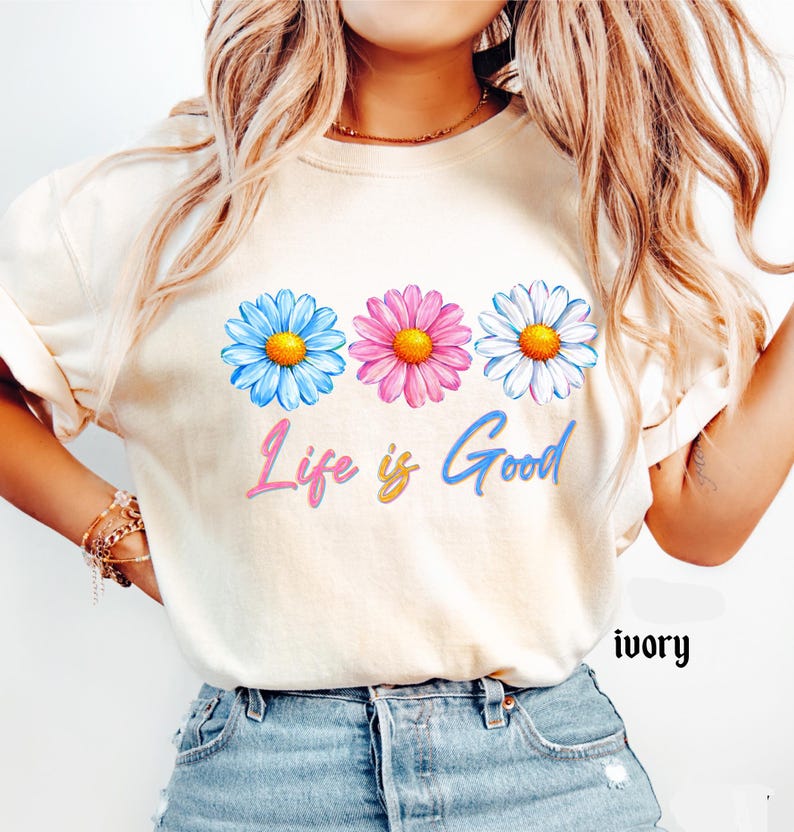Pode incluir: Camiseta cor marfim com tr&ecirc;s margaridas azuis, rosas e brancas com centros amarelos. A frase "Life is Good" est&aacute; escrita abaixo das flores em um gradiente de rosa, amarelo e azul.