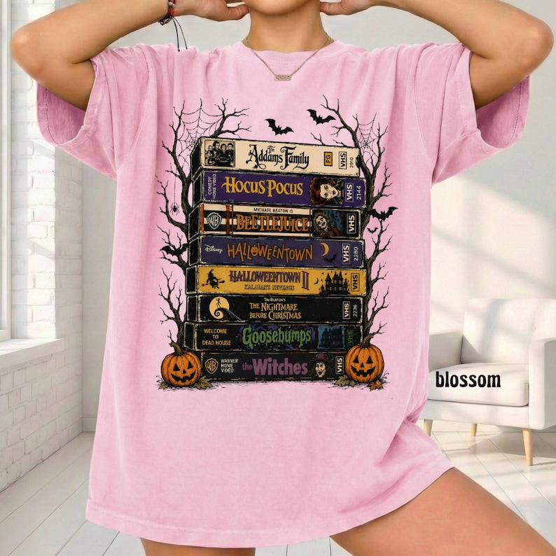 Op de afbeelding: Een lichtroze t-shirt met een grafische print van een stapel van zeven vintage VHS-banden met de labels The Addams Family, Hocus Pocus, Beetlejuice, Halloweentown, Halloweentown II, The Nightmare Before Christmas, Goosebumps en The Witches.