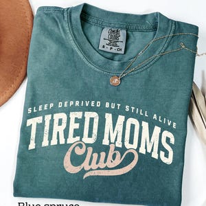 May include: Blue spruce Comfort Colors t-shirt with the text "SLEEP DEPRIVED BUT STILL ALIVE TIRED MOMS Club" in white and pink lettering. A gold necklace with a circular pendant rests on the shirt. A brown hat is visible in the upper left corner.