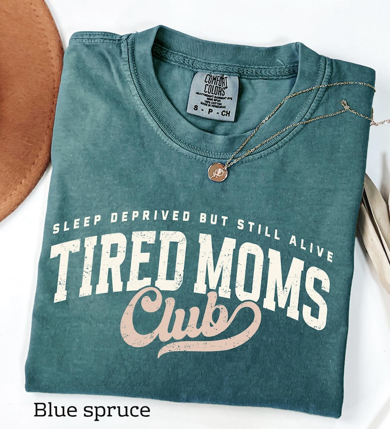 May include: Blue spruce Comfort Colors t-shirt with the text "SLEEP DEPRIVED BUT STILL ALIVE TIRED MOMS Club" in white and pink lettering. A gold necklace with a circular pendant rests on the shirt. A brown hat is visible in the upper left corner.