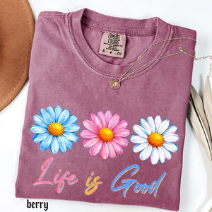 Pode incluir: Camiseta cor de berry com tr&ecirc;s flores de margarida em azul, rosa e branco, cada uma com um centro amarelo. A frase "Life is Good" est&aacute; escrita em letras coloridas abaixo das flores. Um colar de ouro com um pingente repousa sobre a camisa.