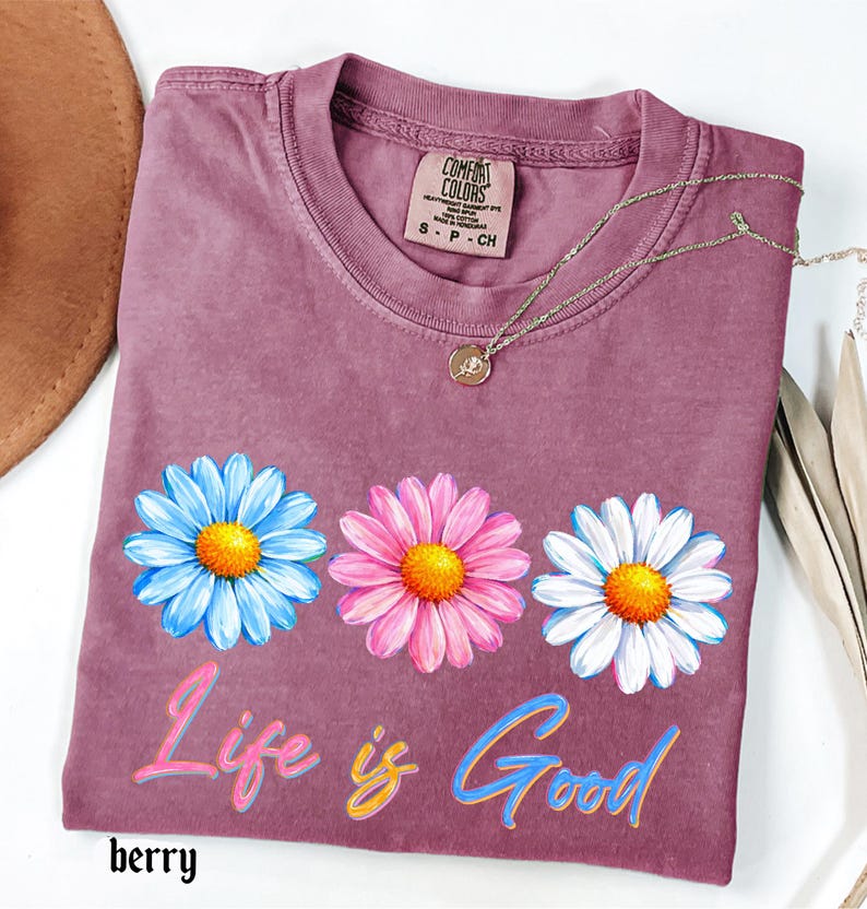 Pode incluir: Camiseta cor de berry com tr&ecirc;s flores de margarida em azul, rosa e branco, cada uma com um centro amarelo. A frase "Life is Good" est&aacute; escrita em letras coloridas abaixo das flores. Um colar de ouro com um pingente repousa sobre a camisa.