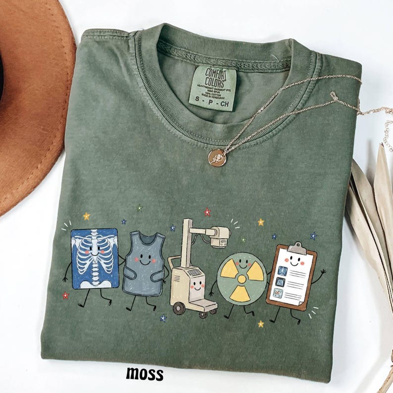 May include: A moss green crew neck t-shirt featuring a graphic print of five anthropomorphic medical items including a skeleton x-ray, a lead apron, an x-ray machine, a radiation symbol, and a clipboard, all with smiling faces and small stars.