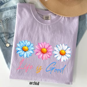 Pode incluir: Camiseta cor de orqu&iacute;dea com tr&ecirc;s flores de margarida em azul, rosa e branco, com centros amarelos. O texto "Life is Good" est&aacute; impresso abaixo das flores em rosa e azul. A palavra "orchid" est&aacute; impressa na parte inferior.