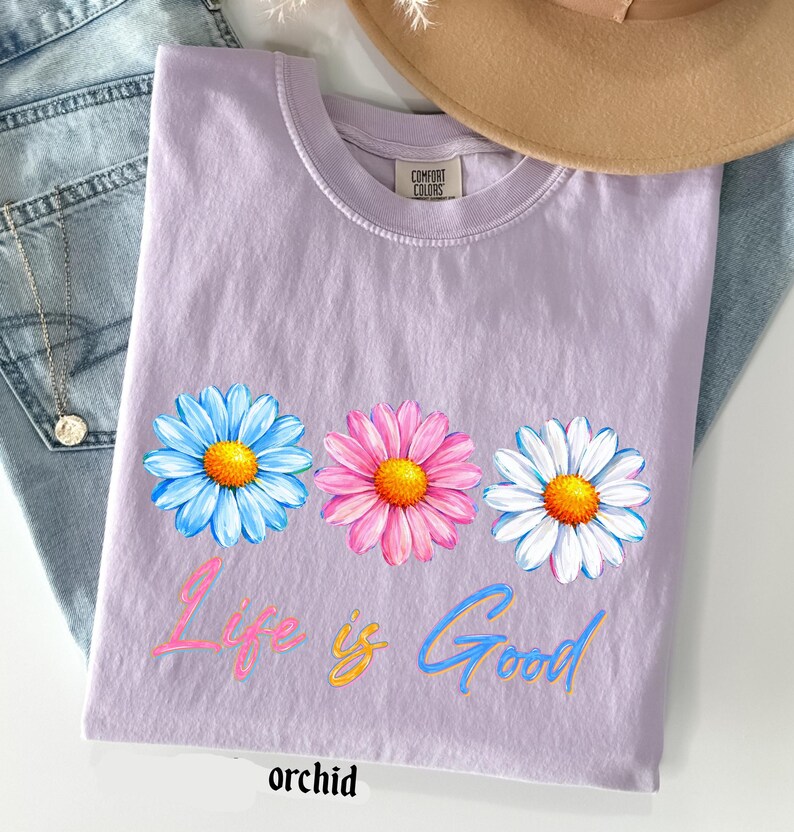 Pode incluir: Camiseta cor de orqu&iacute;dea com tr&ecirc;s flores de margarida em azul, rosa e branco, com centros amarelos. O texto "Life is Good" est&aacute; impresso abaixo das flores em rosa e azul. A palavra "orchid" est&aacute; impressa na parte inferior.