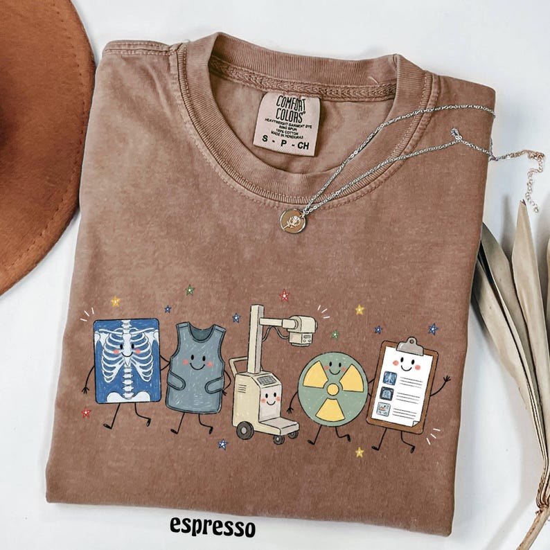 May include: A brown short-sleeve t-shirt featuring a graphic of five cartoon medical imaging items including an X-ray, a lead apron, an imaging machine, a radiation symbol, and a clipboard. The shirt has a Comfort Colors label in the neck.