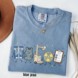 May include: A blue jean colored t-shirt with a crew neckline and a Comfort Colors brand label. The graphic features cartoon illustrations of a chest x-ray, a lead apron, an x-ray machine, a radiation symbol, and a clipboard, all with smiling faces.